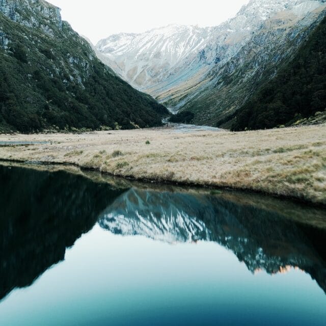 Drone view of a remote Southern Alps valley during a free-range chamois hunt in New Zealand with mountain reflections in calm water.